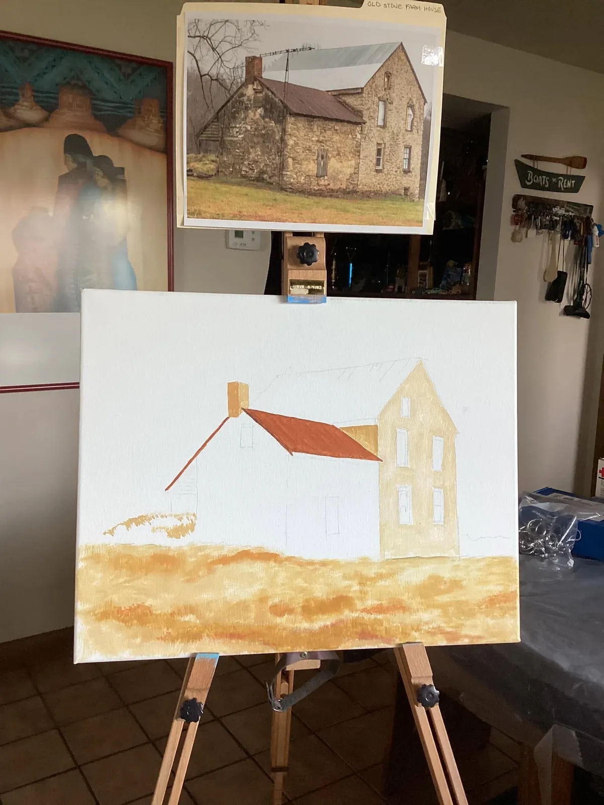An unfinished oil painting of a stone farmhouse on a wooden easel, with the reference photograph clipped above it